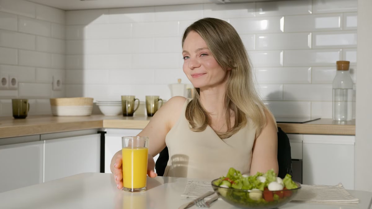 Smiling woman enjoying a healthy meal with salad and orange juice in a modern kitchen.